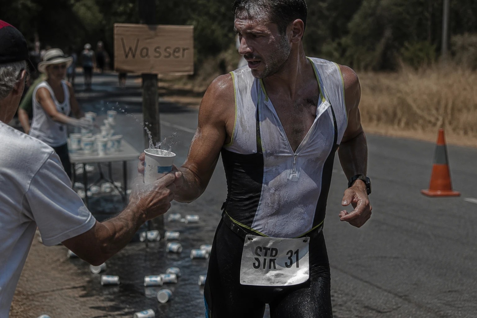 Triathlet greift bei praller Mittagssonne an einer Verpflegungsstation nach einem Wasserbecher, Salzränder auf Schläfen und Trikot, dahinter ein Schild mit der Aufschrift Wasser.