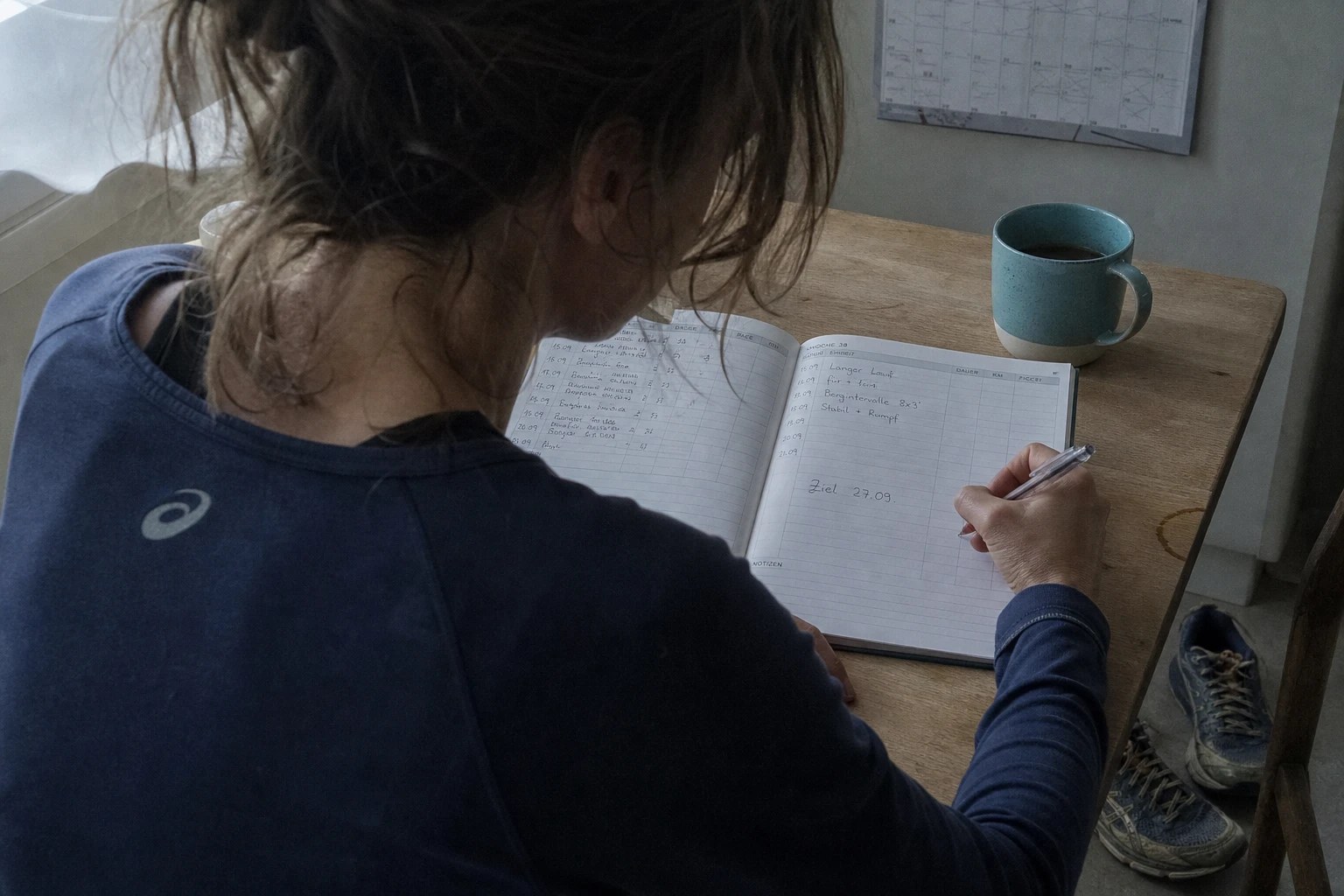 Läuferin sitzt frühmorgens am Küchentisch und trägt von Hand ein Renndatum in ein Trainingstagebuch ein, daneben abgetragene Laufschuhe und eine Tasse.